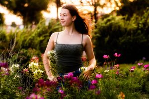 Young woman with flowing hair smiles among colorful garden flowers at golden hour sunset.