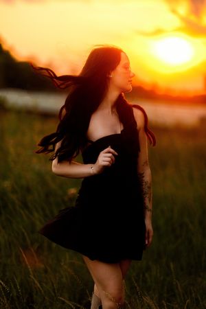 Woman in black dress standing in golden sunset field, long dark hair blowing in wind, tattoos visible.