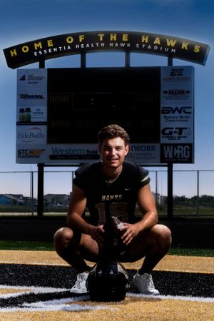 Baseball player squatting in front of stadium scoreboard on sunny day.