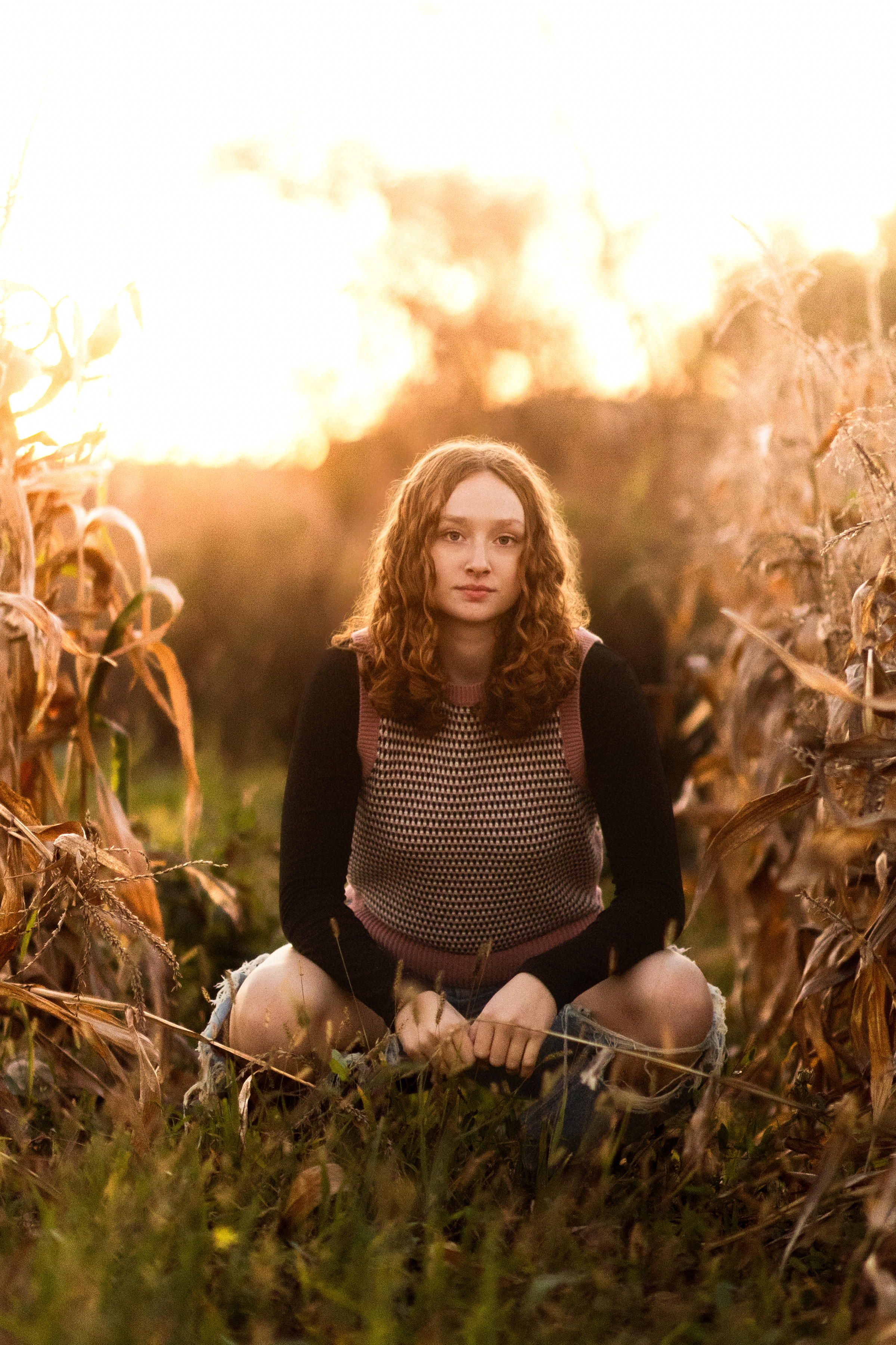 A person wearing a brown sweater sits among tall dried corn stalks at sunset in a rustic autumn field.