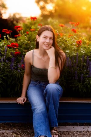 Young woman sitting in a garden at golden hour, smiling with red flowers and purple blooms behind her.