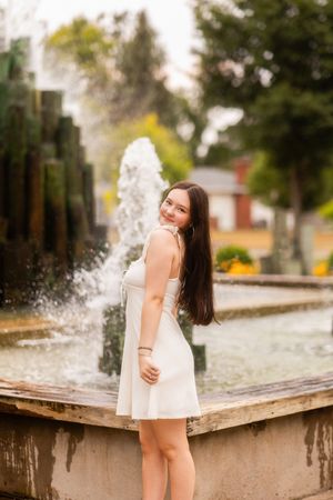 Young woman in white sundress smiling by a park fountain with lush green trees in the background.