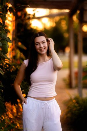 Young woman in pink top and white pants smiling outdoors at golden hour sunset in a garden setting.