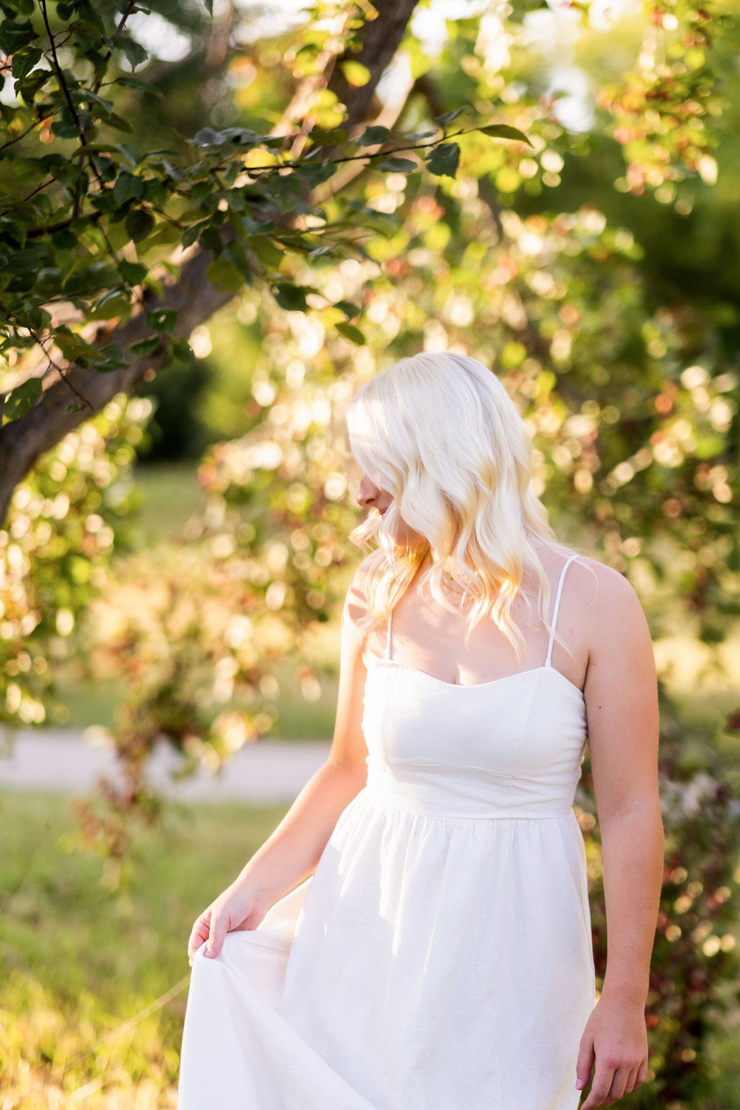 Blonde woman in white sundress standing under sunlit green tree branches in an outdoor garden setting.