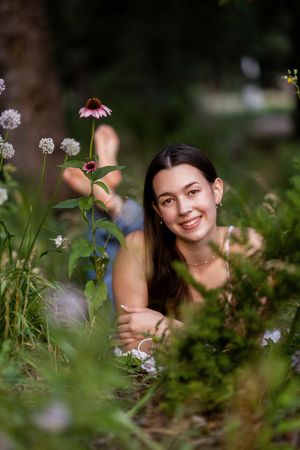 A smiling young woman with long dark hair lying among lush green garden plants and pink echinacea flowers outdoors.