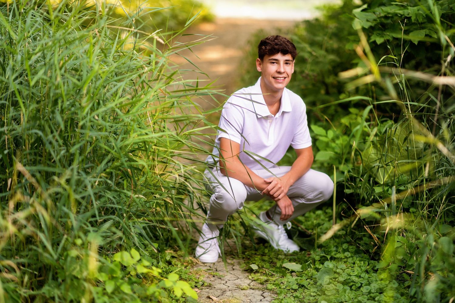Young man in white polo and gray pants crouching on a stone path surrounded by lush green plants outdoors.