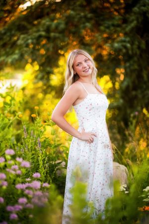 Smiling blonde woman in white floral sundress standing among colorful garden flowers in golden hour sunlight.