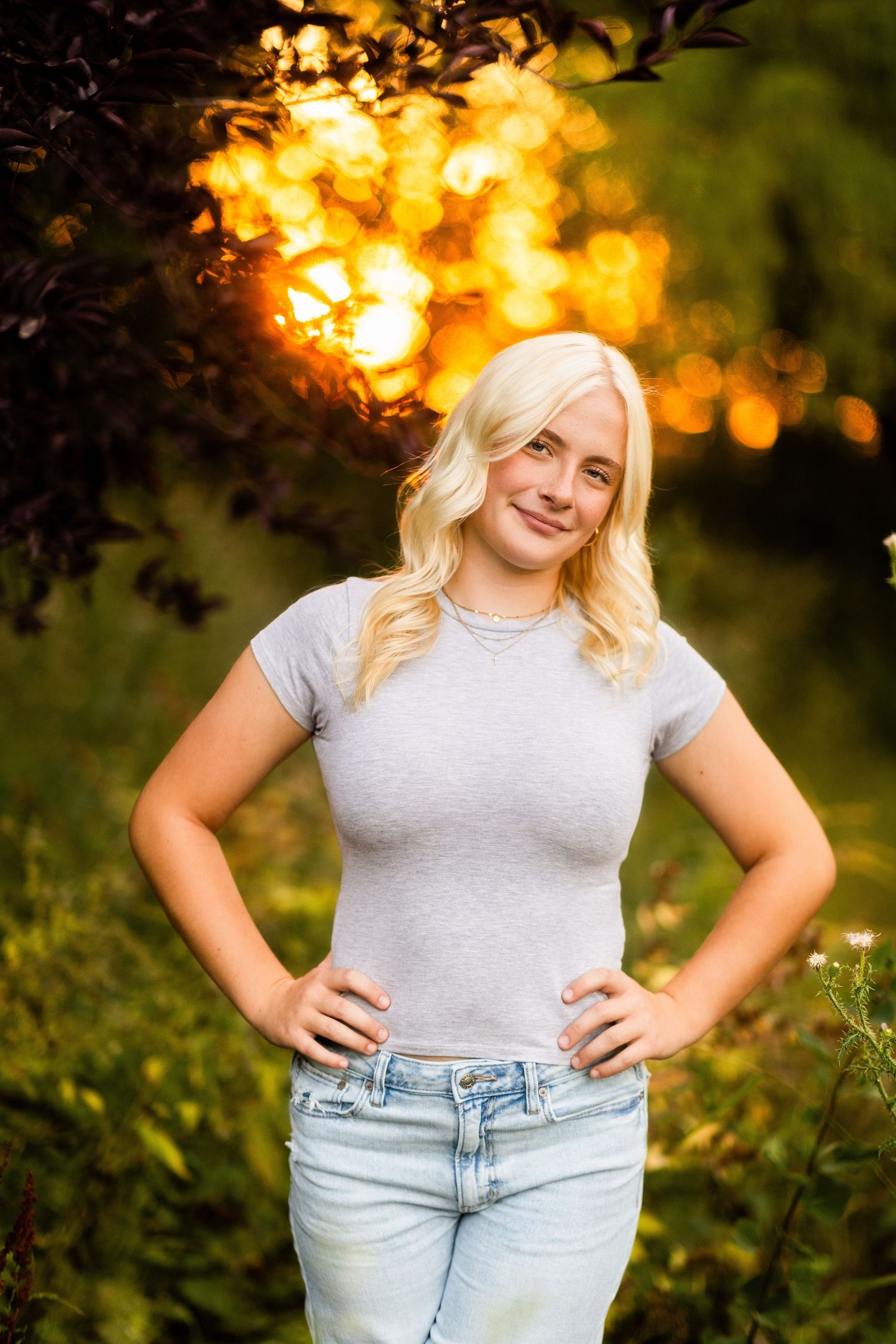 Blonde woman in gray t-shirt and jeans poses outdoors with golden sunset bokeh light behind her.