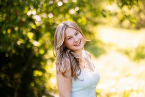 A woman in a white dress poses outdoors in golden sunlight with trees and bokeh lighting effects in the background.