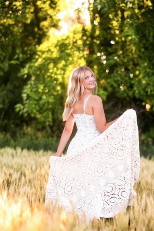Blonde woman in white lace maxi dress holding skirt in golden wheat field with green trees at sunset.