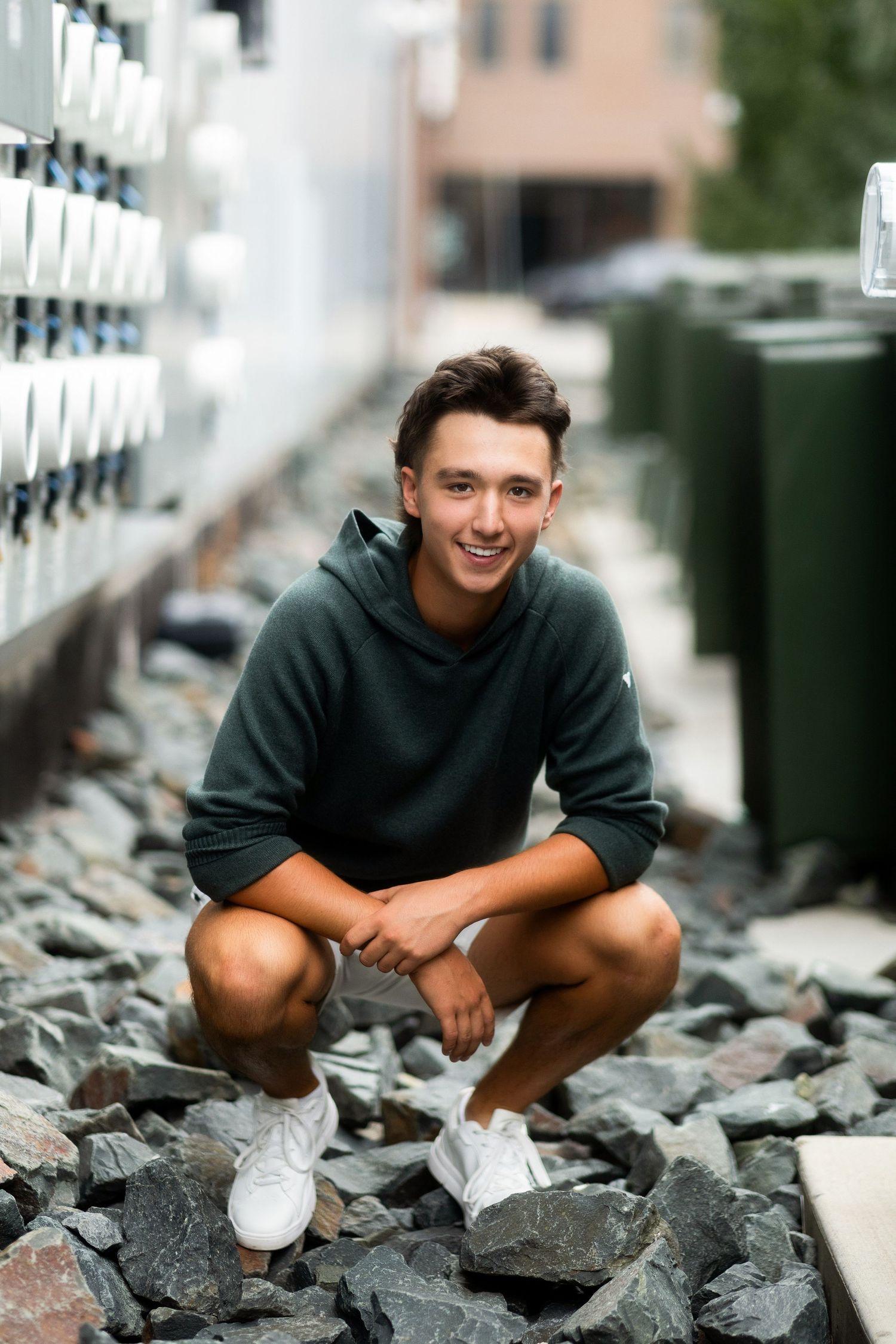 Young man in green hoodie and shorts crouching on rocks, smiling confidently in an urban outdoor setting.