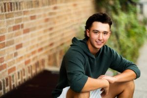 Young man in green hoodie smiling while sitting against a brick wall outdoors.
