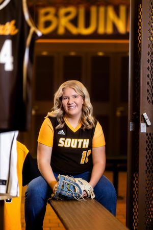 Student in yellow and black athletic uniform sits on blue bench in front of Bruin sign for senior portraits.