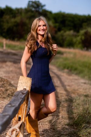 Smiling blonde woman in navy blue mini dress and tan boots posing by a rustic wooden fence in a sunny rural field.