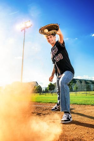 Young baseball player in black jersey number 20 poses with glove raised on a dusty field under bright stadium lights.
