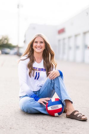Smiling female volleyball player sitting on pavement holding a Tachikara ball, wearing a white team jersey and jeans.