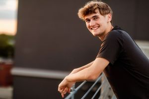 Smiling young man in black t-shirt leaning on railing outdoors at sunset with urban background.