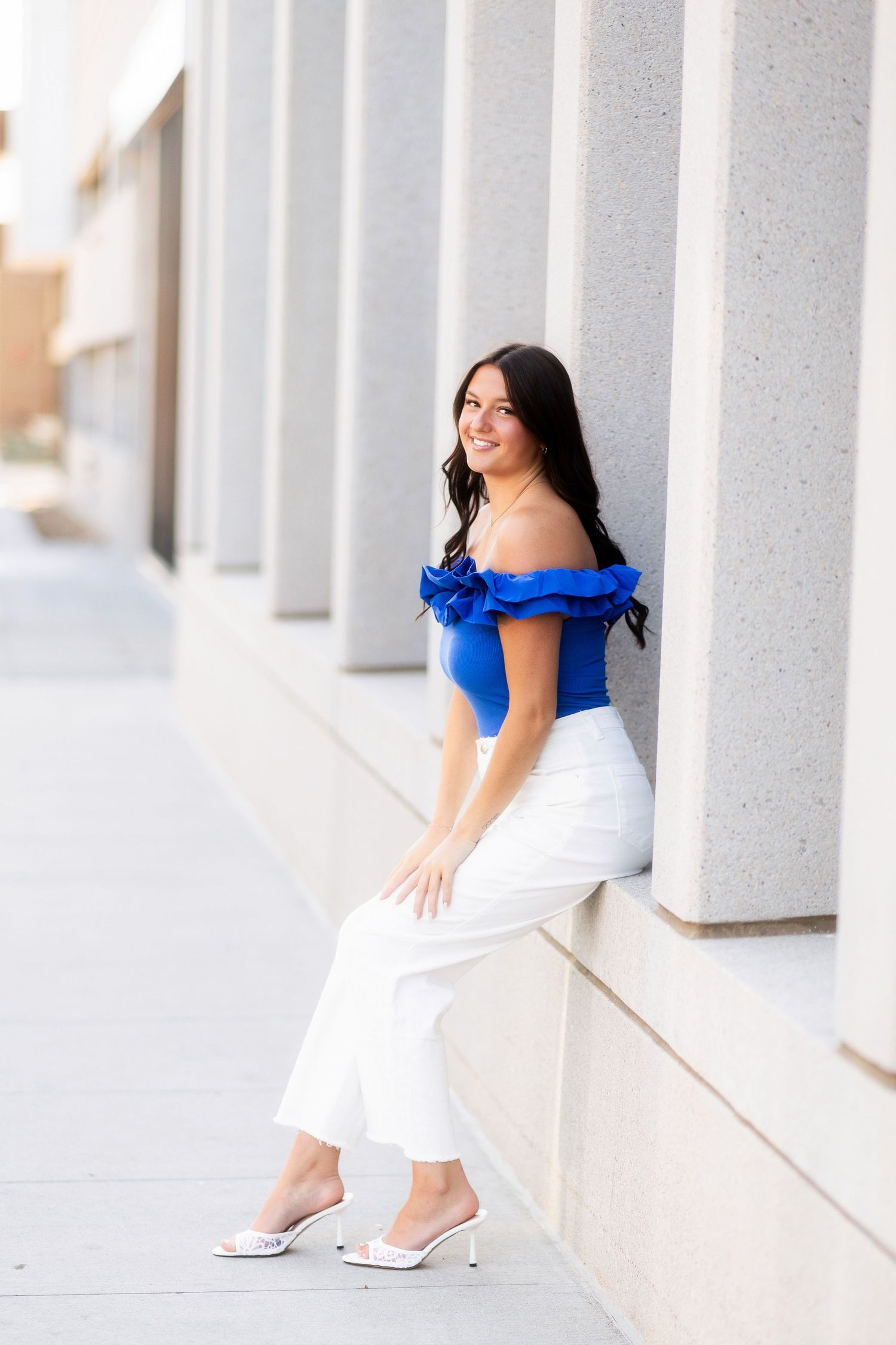 Young woman in blue off-shoulder ruffled top and white pants poses against stone columns outdoors.