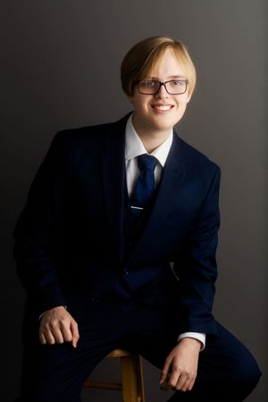 Professional studio portrait of someone wearing a navy blue suit, white dress shirt, tie and glasses against dark background.