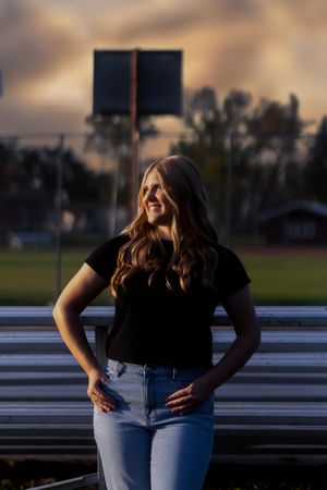 A person in a black t-shirt and jeans poses outdoors at sunset with bleachers in the background.