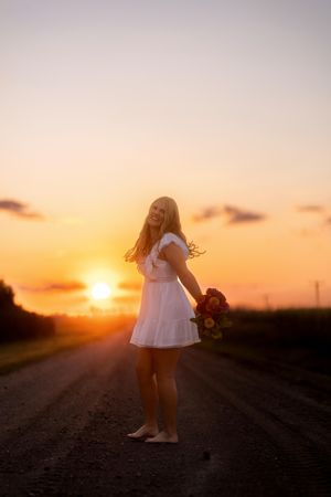 A silhouette of a person in a white dress stands on a dirt road during a golden sunset.