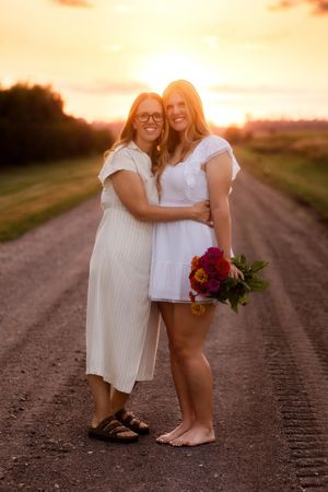 Two people share a joyful embrace on a dirt road at sunset while holding a vibrant red bouquet.