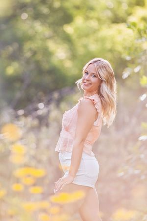Smiling blonde woman in pink top and white shorts standing in a sunlit field with yellow wildflowers.