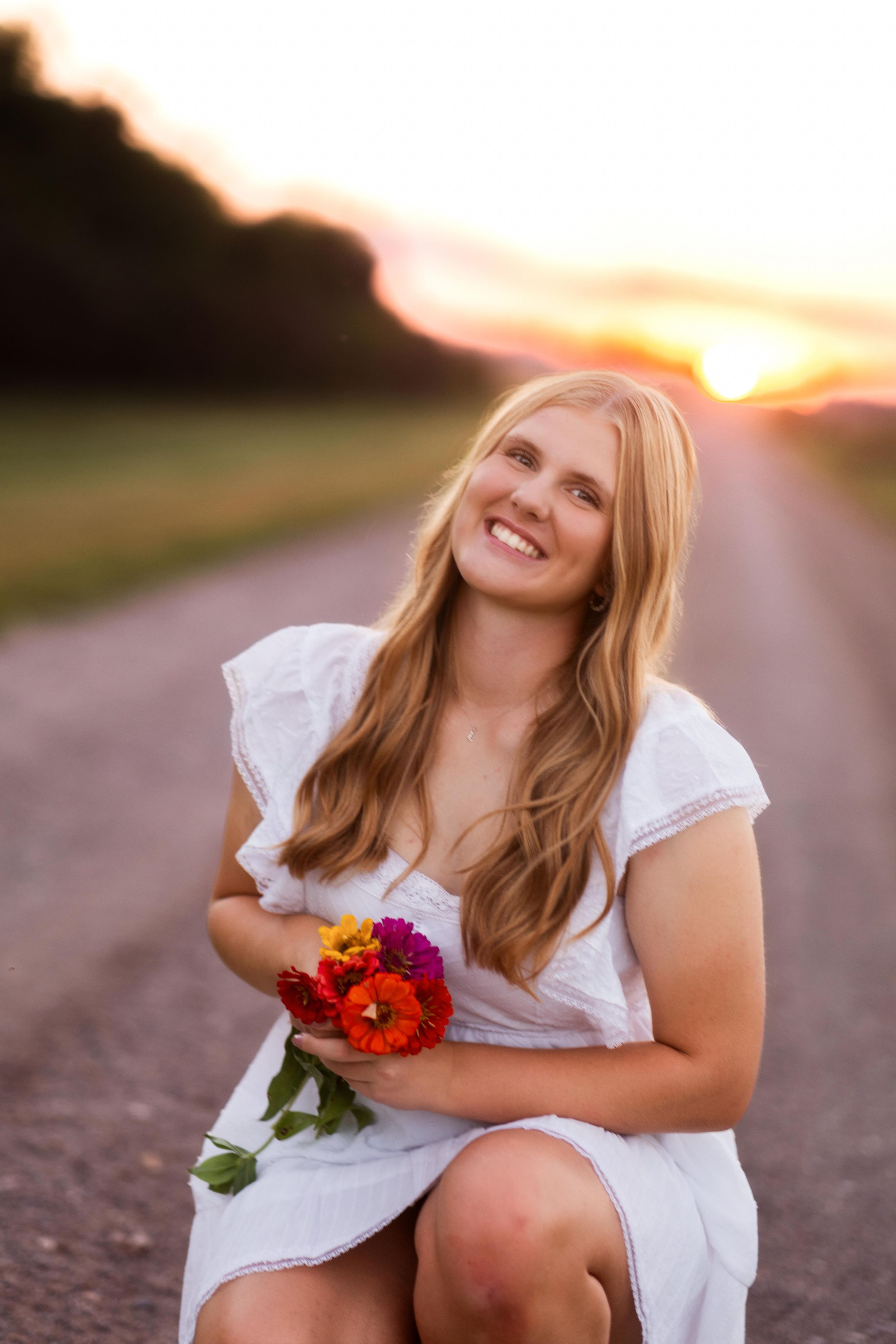 A person in a white dress poses with colorful flowers against a sunset backdrop on a country road.