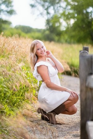 Smiling blonde woman in white dress and brown boots crouching by a rustic fence in a sunny field.