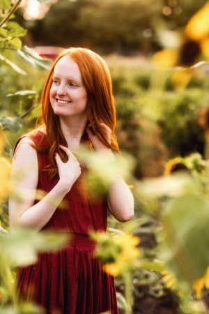 A smiling young woman with long red hair in a burgundy dress poses among sunflowers in a golden garden.
