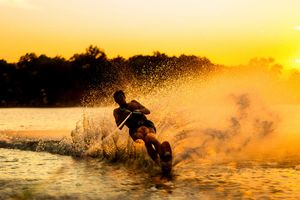 Person water skiing at golden sunset, creating dramatic water spray on a calm lake with silhouetted trees in background.