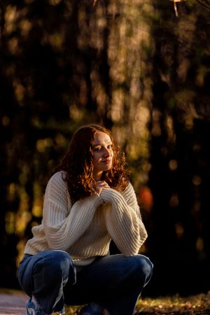 Person in white sweater sitting thoughtfully on grass during fall season with blurred forest background.