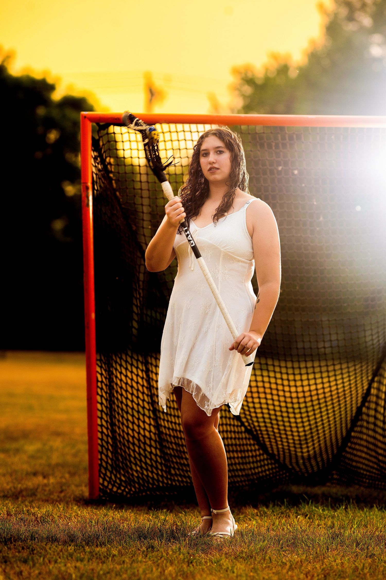 Young woman in white dress holding lacrosse stick stands before a red goal net at golden hour sunset.