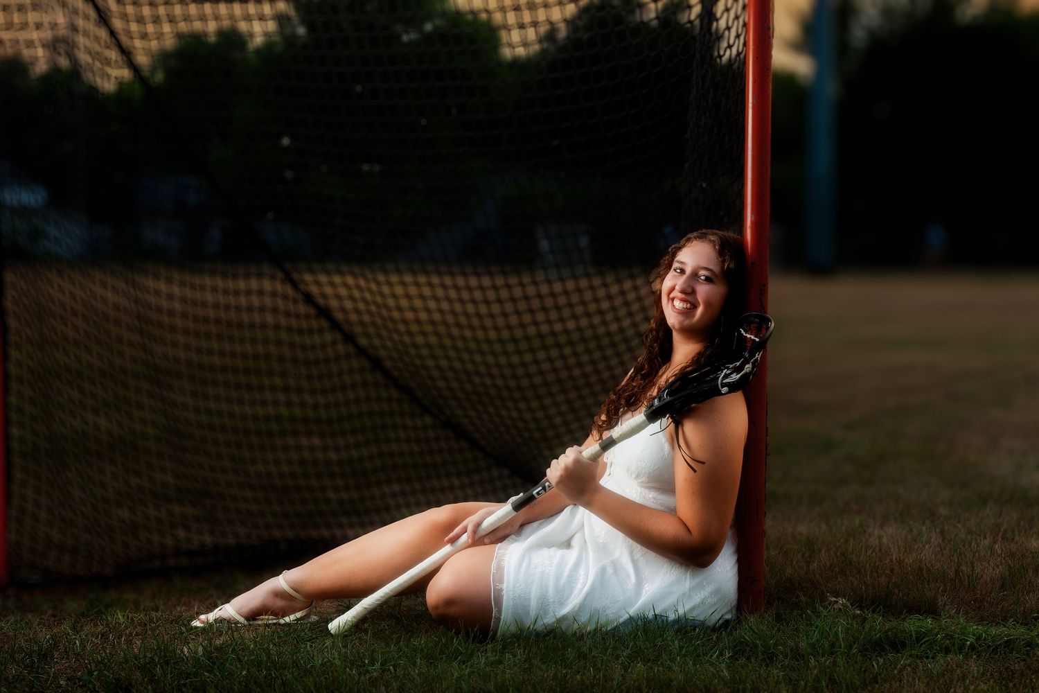 Young woman in white dress sitting by lacrosse goal post on grass field, holding lacrosse sticks, smiling at sunset.