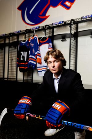 Hockey player sits in locker room wearing blue and orange team jersey and gloves against dark background.
