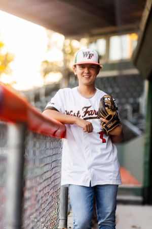 A baseball player in a white uniform and cap leans against a dugout fence with a glove.