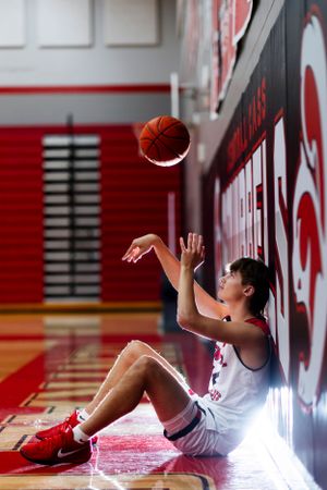 Basketball player sits against gym wall tossing ball upward in dramatic lighting.
