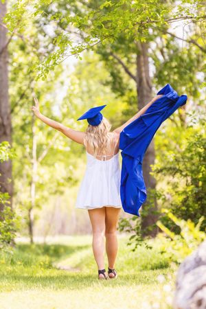 A person in a blue graduation gown twirls joyfully outdoors in a sunny park setting with green trees in the background.
