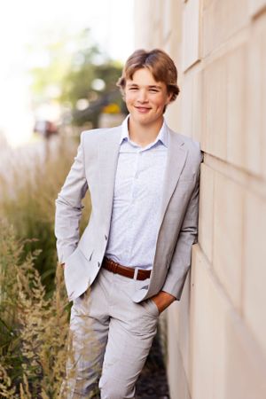 Figure in light gray suit and white shirt posing against white wall outdoors.