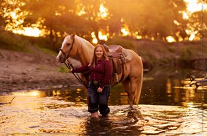 Horse and rider stand in a stream during golden sunset light, creating a magical outdoor scene.