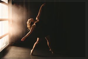Silhouetted dancers perform expressive movements against dramatic window lighting in a dark studio space.