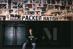 Person wearing a Green Bay Packers jersey sits beneath a wall covered in team memorabilia and 'Packer Nation' signage.