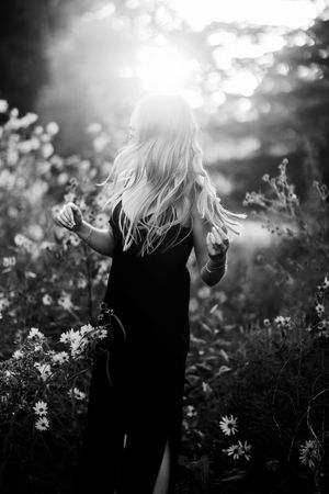 Silhouette of a person in a long dress standing among wildflowers in sunlit field, captured in black and white.