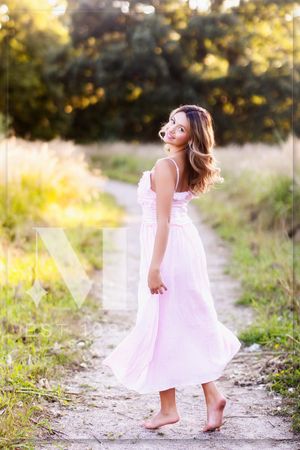 A barefoot person in a flowing light pink dress twirls on a sunlit outdoor path during golden hour.