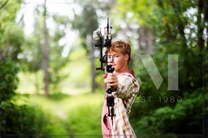An archer in plaid shirt draws back a compound bow while practicing in a sunlit forest setting.