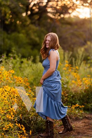 Figure in a flowing blue dress poses with wildflowers at golden hour, with warm sunlight filtering through trees.