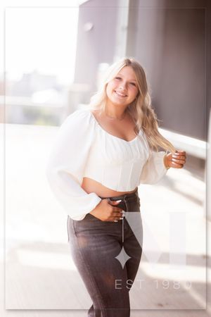 Fashion model poses in white top and dark pants against bright window backdrop.