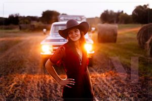 Woman in red dress and cowboy hat poses in front of hay bales at sunset.