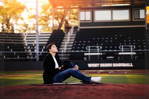 A person in black attire sits on a baseball field during golden hour with empty stadium seats in the background.
