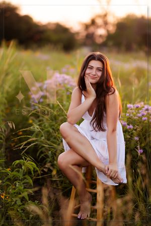A person in a white summer dress sits barefoot among purple wildflowers during golden hour.
